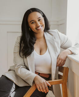 A woman with long dark hair sits in a wooden chair by a window, smiling warmly. She wears a light beige blazer over a white top.”