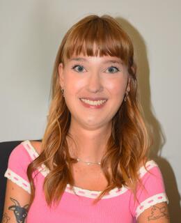 Photo of Erin smiling at the camera. She is a white woman with brown hair, silver jewelry and piercings, wearing a pink shirt.