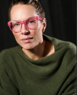 A middle aged white woman wearing pink glasses and an olive green sweater is pensively smiling at the camera