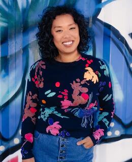 A Filipina woman with shoulder-length curly black hair is standing and smiling in front of a spray painted mural.